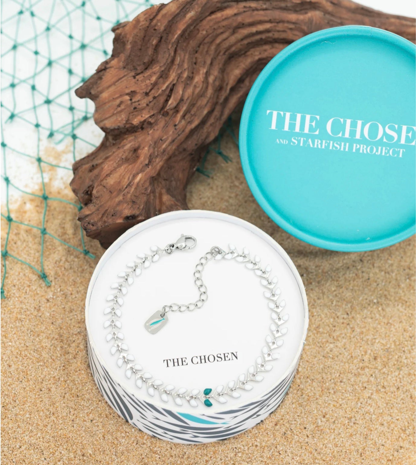 Silver bracelet on a card with 'The Chosen' branding, next to driftwood and a turquoise container on sand.