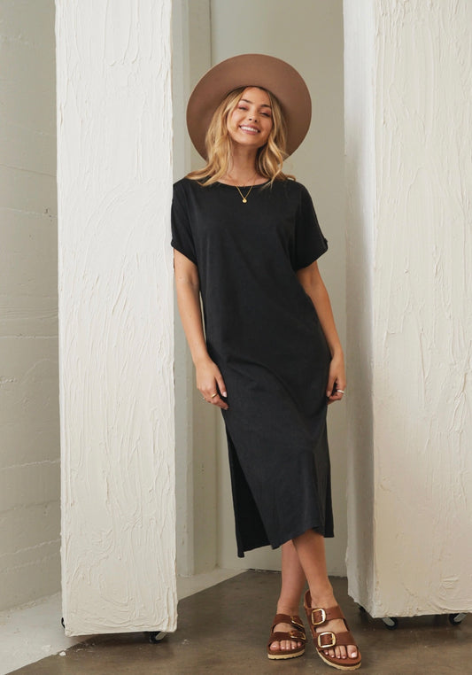 Woman wearing a black dress and brown hat standing against a white wall.