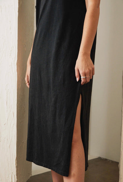 Person wearing a black dress with a slit, standing against a neutral wall.